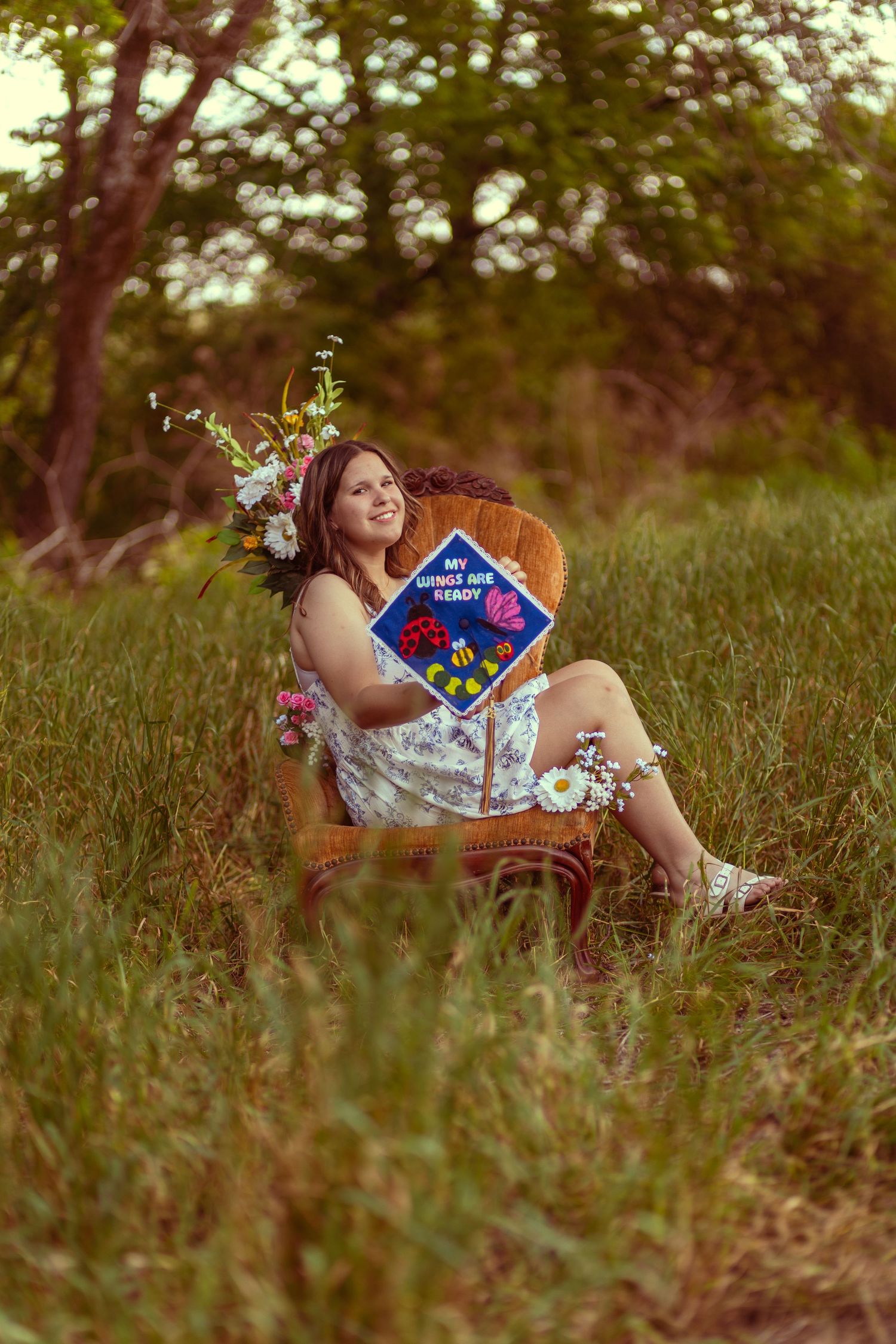 Young woman in floral dress sitting in wicker chair in meadow, holding graduation cap with flower crown.