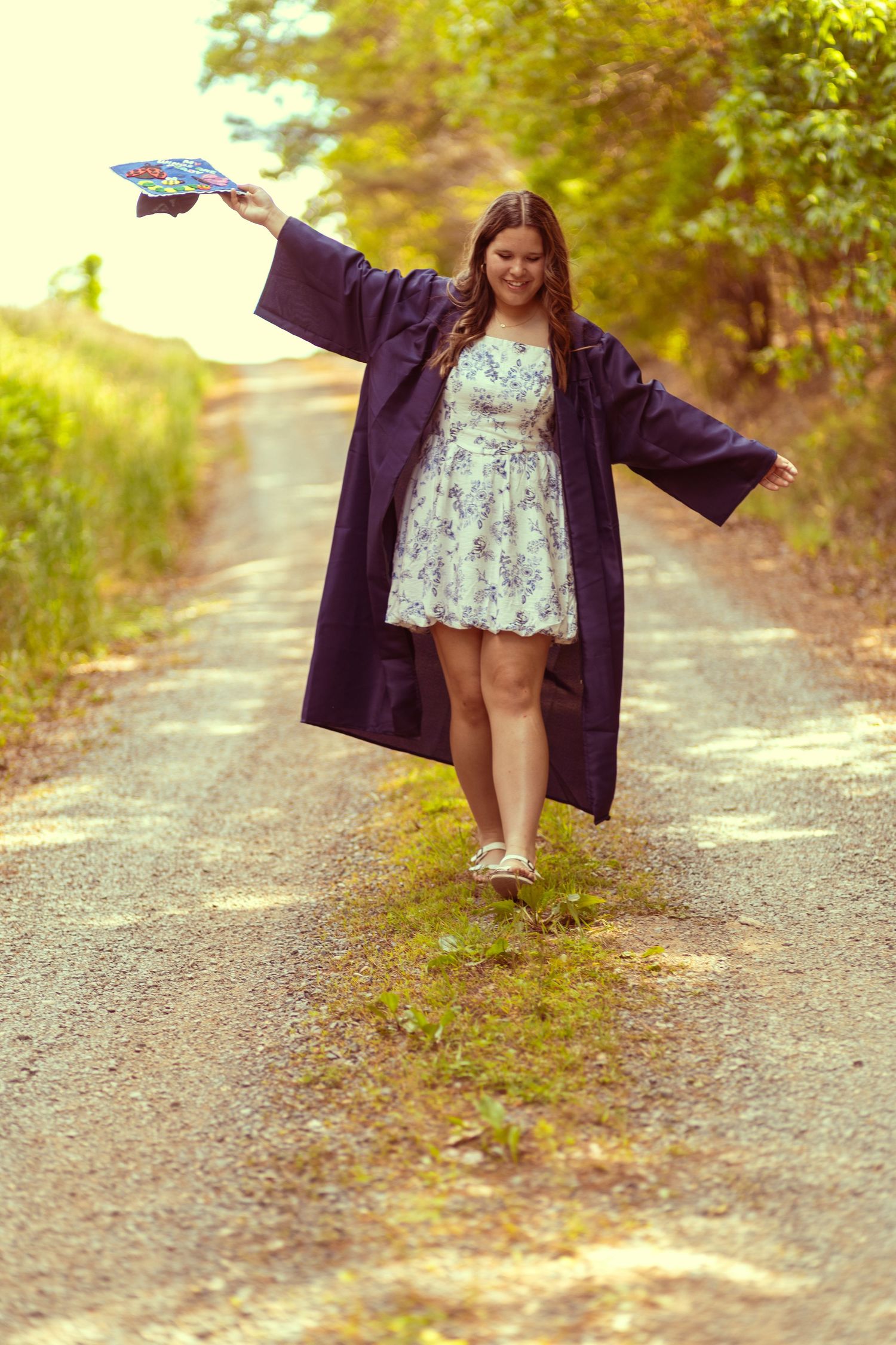 Young woman in graduation gown and floral dress celebrating on a rural road, holding cap aloft with arms outstretched.