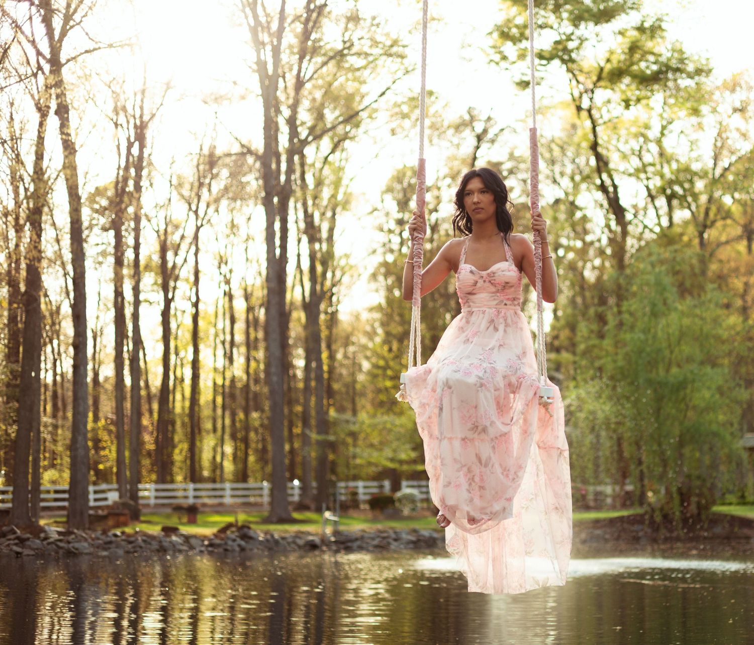 A woman in a flowing pink floral gown sits on a rope swing over water surrounded by sunlit trees.
