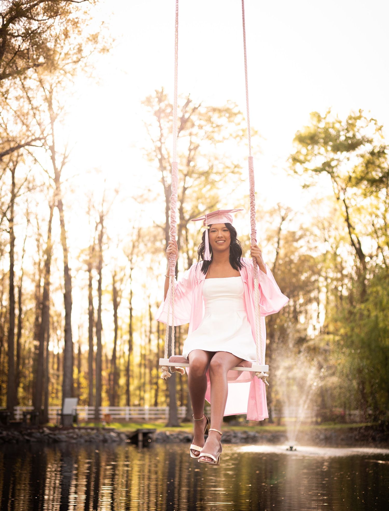 Graduate in pink cap and gown sits on rope swing over pond surrounded by sunlit trees.