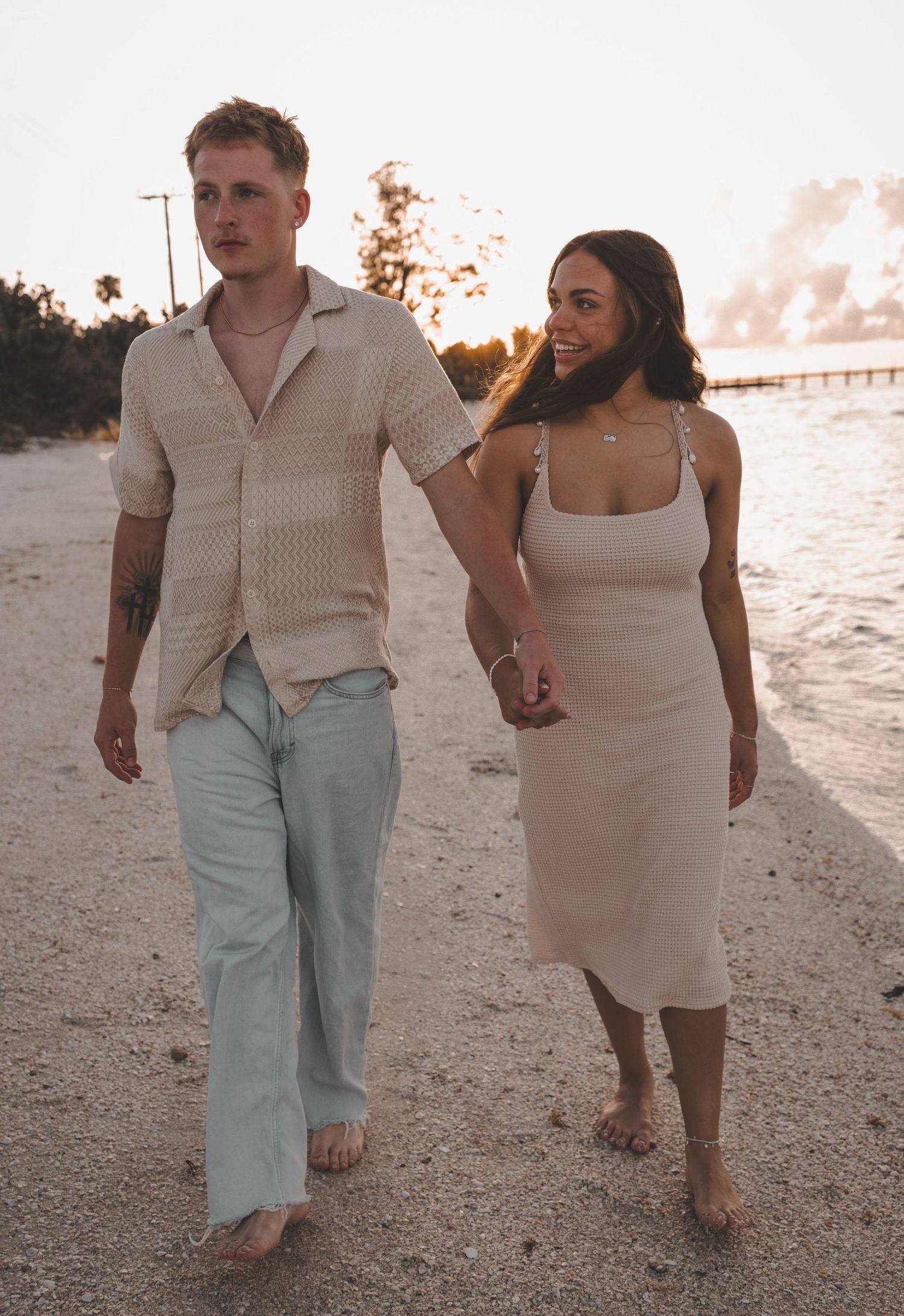 Couple walking hand in hand on beach at sunset in summer attire.
