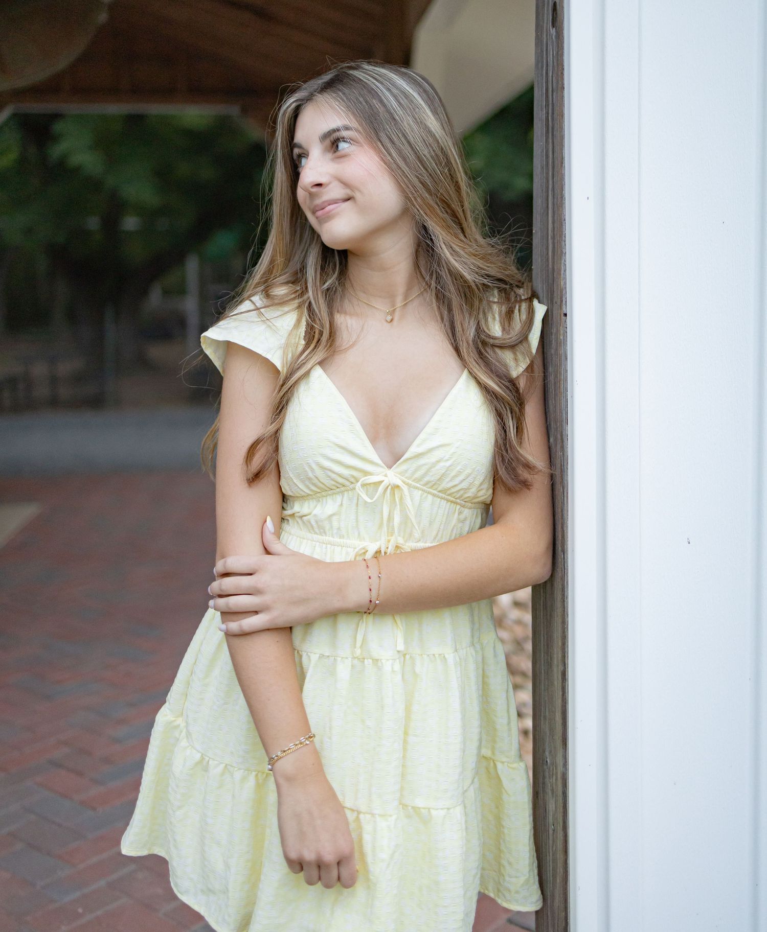 Young woman in a light yellow mini dress leaning against a white pillar, smiling outdoors.