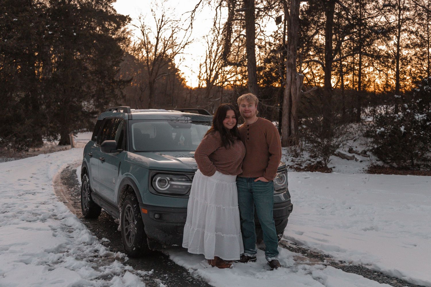 Couple smiling together beside a teal Ford Bronco Sport on a snowy road at sunset in winter.