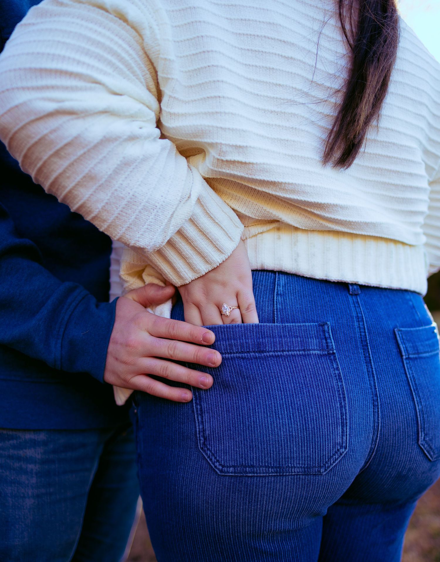 Couple embracing from behind, woman in cream sweater and blue jeans with a ring on her finger.