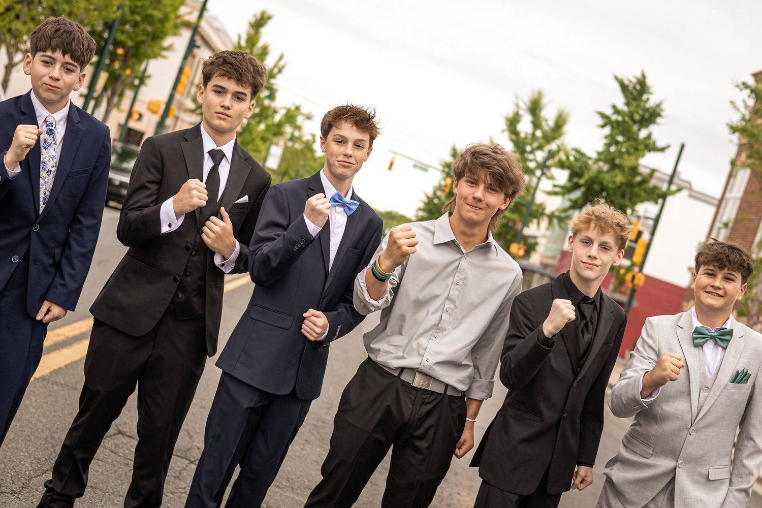 Six teenage boys in formal suits and ties pose with fists raised on a street, dressed for a special occasion.