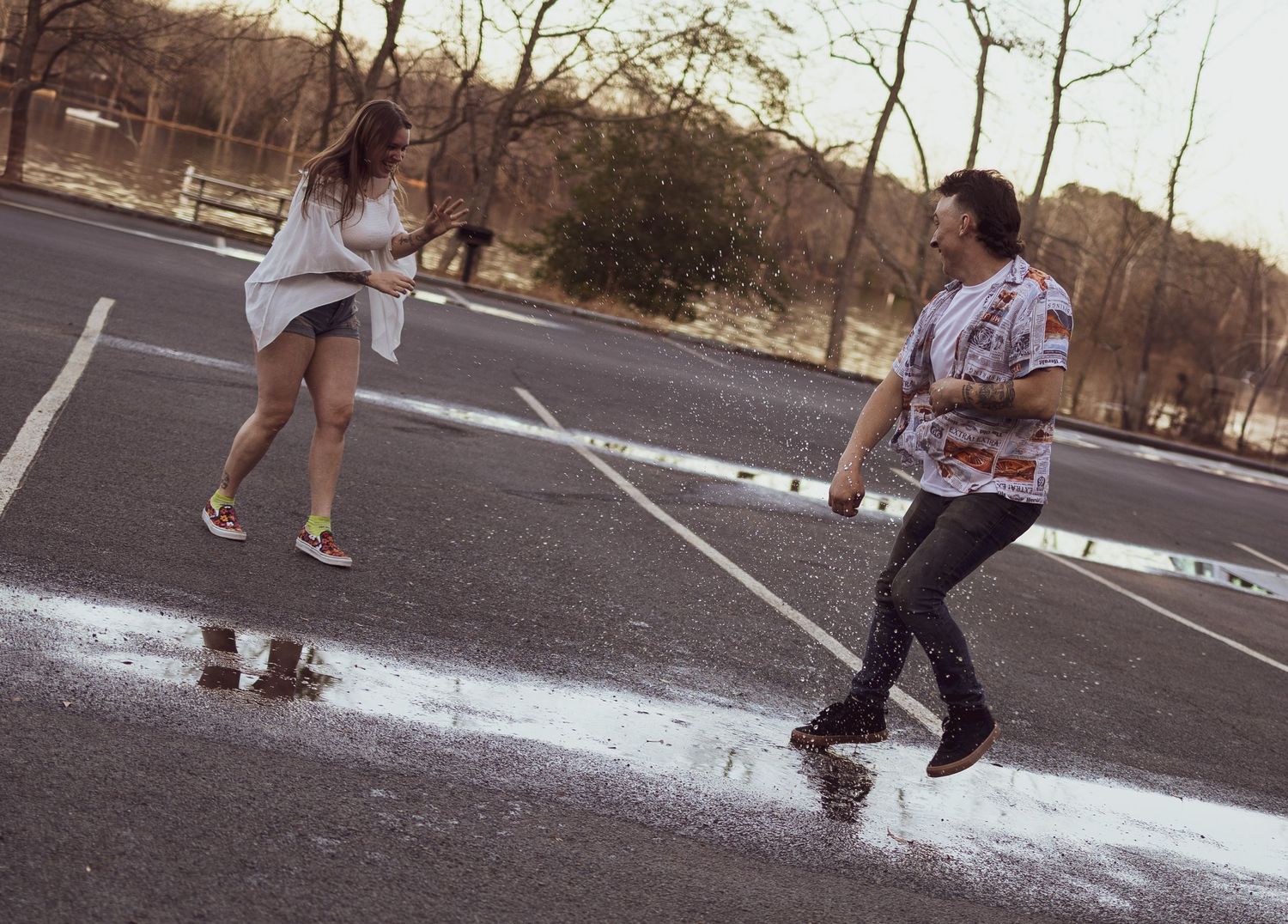 Two people dancing playfully in a wet parking lot surrounded by bare trees on a cloudy day.