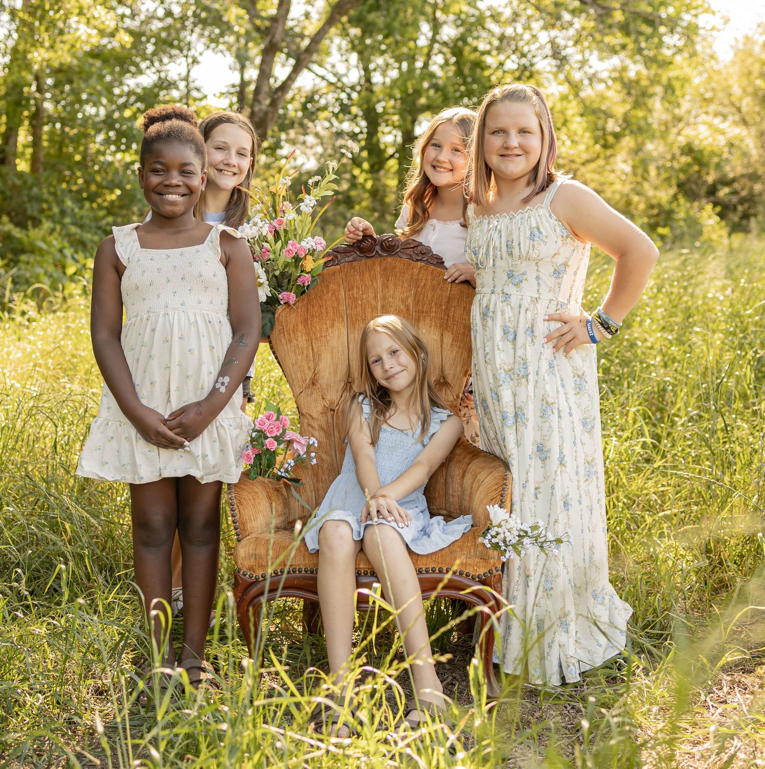 Five young girls pose together in a sunlit meadow, wearing floral dresses, with one seated in a vintage wicker throne chair.