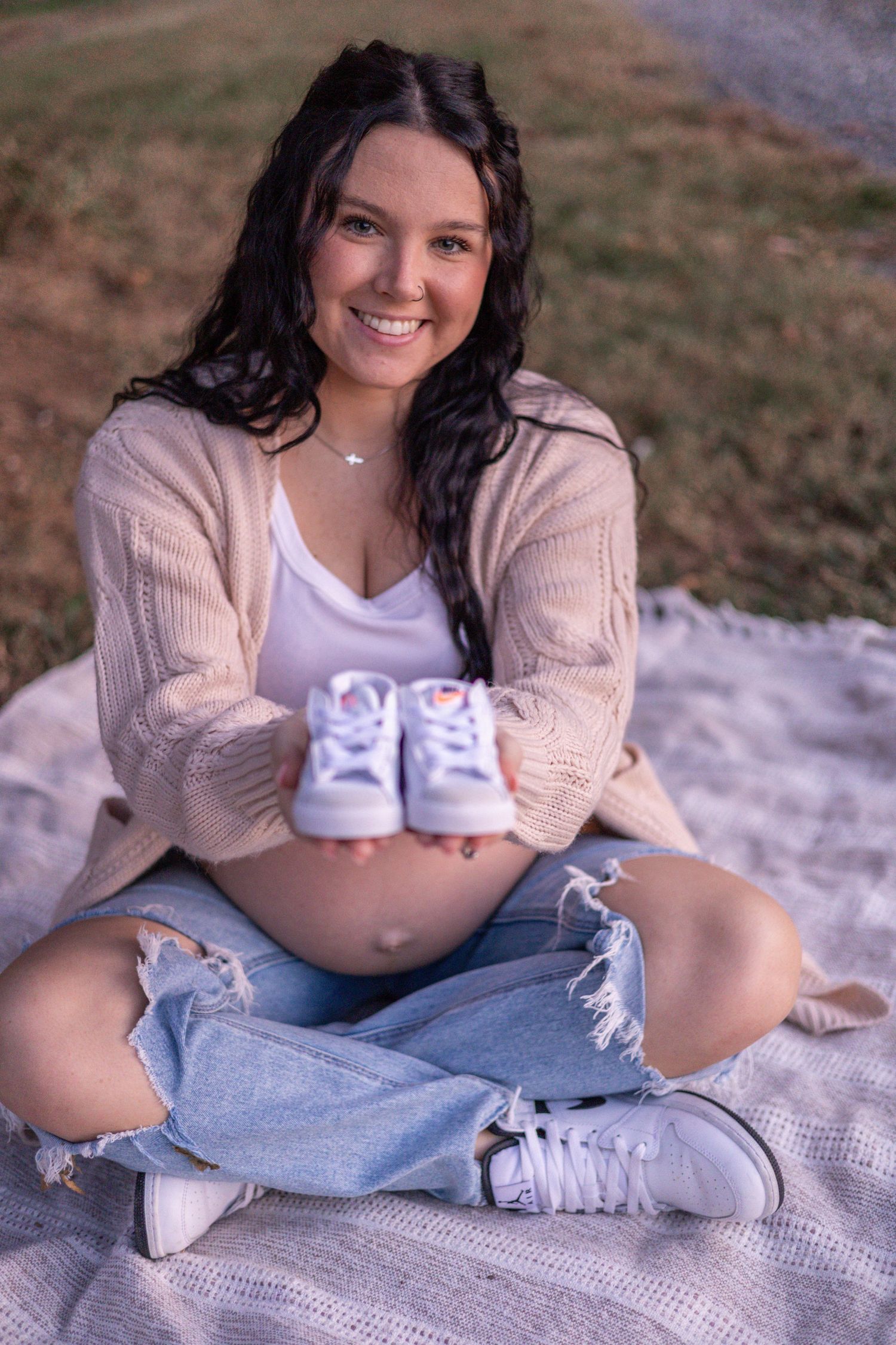 Expectant mother in beige cardigan and ripped jeans sits on blanket outdoors holding tiny baby shoes.