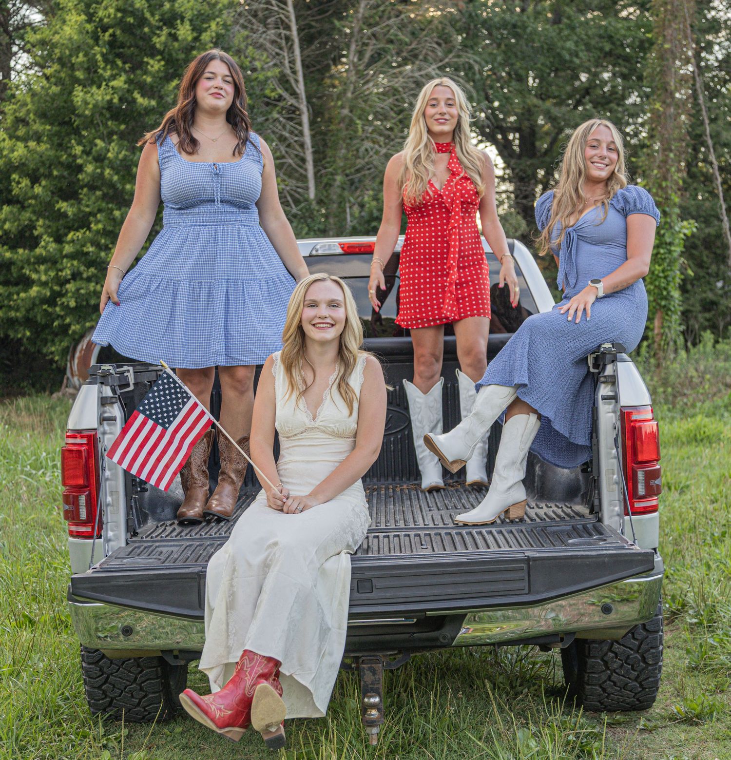 Three friends in western boots sitting casually on the back of a pickup truck in a grassy outdoor setting.
