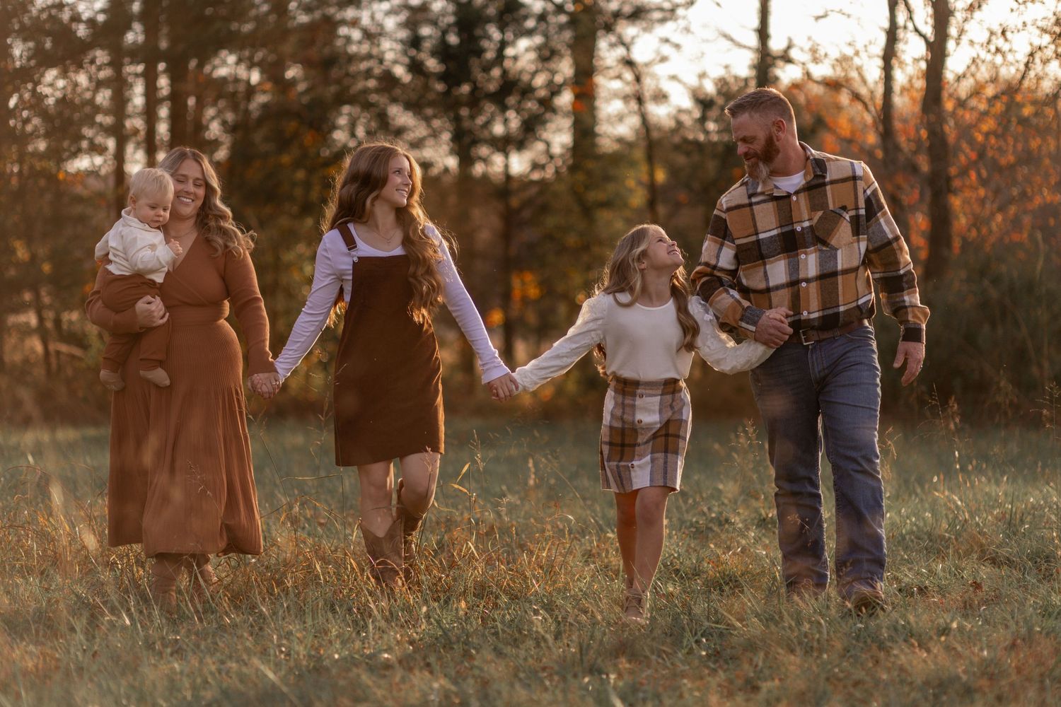A family walks hand in hand through a sunlit meadow at golden hour with trees in the background.