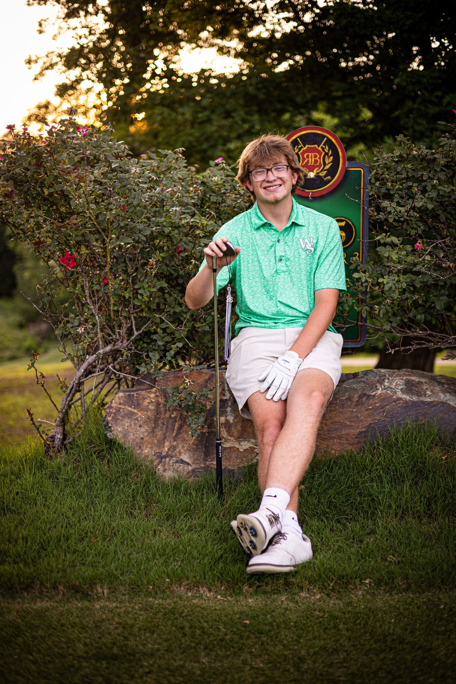 Young golfer in green polo shirt holding a golf club, sitting on rocks near a golf course sign at sunset.