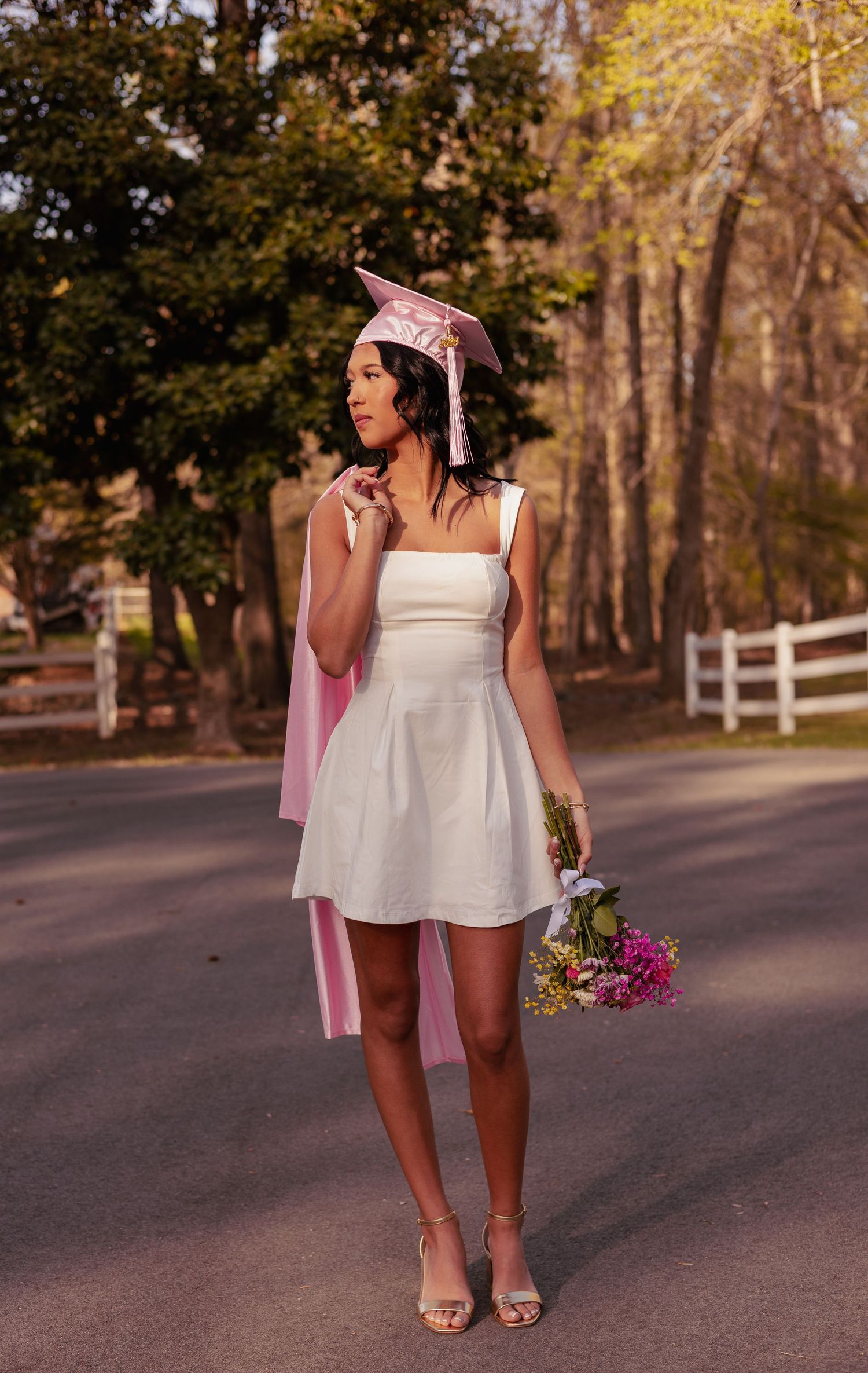 Young woman in white mini dress and pink graduation cap holds flowers on a tree-lined road.