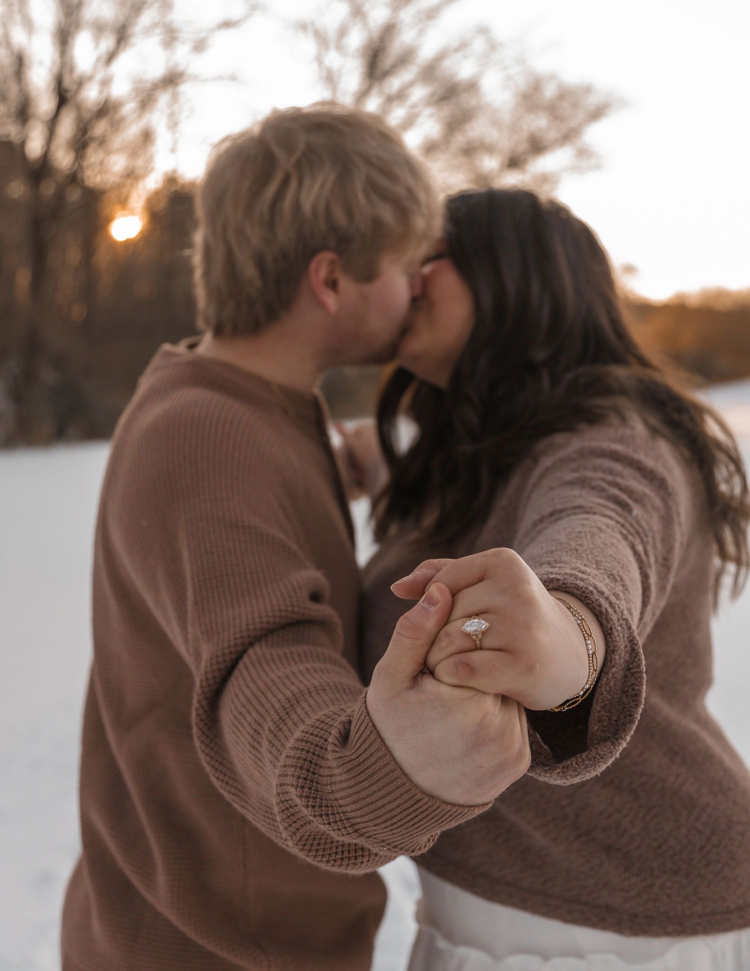 Couple kissing at sunset in snowy field, woman displaying diamond engagement ring while holding hands.