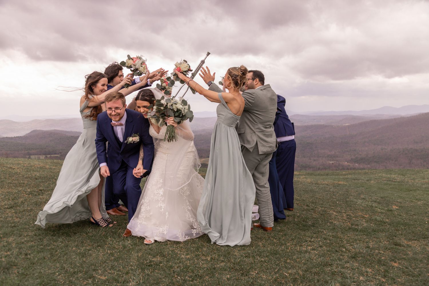 Wedding party celebrates in circle on hilltop with mountain views and overcast sky.