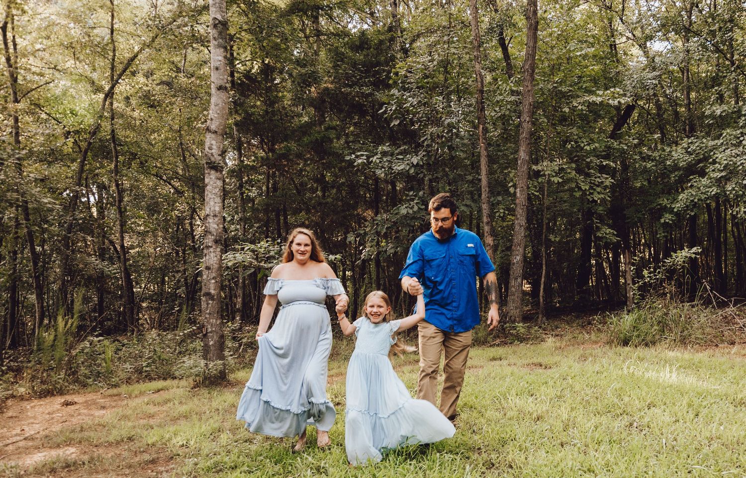 A family in coordinating blue and white outfits poses together in a sunlit forest clearing for outdoor portraits.