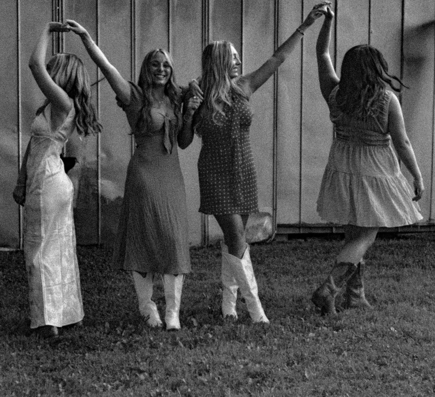 A black and white panoramic photograph shows a row of summer dresses swaying in movement against a wooden fence background.