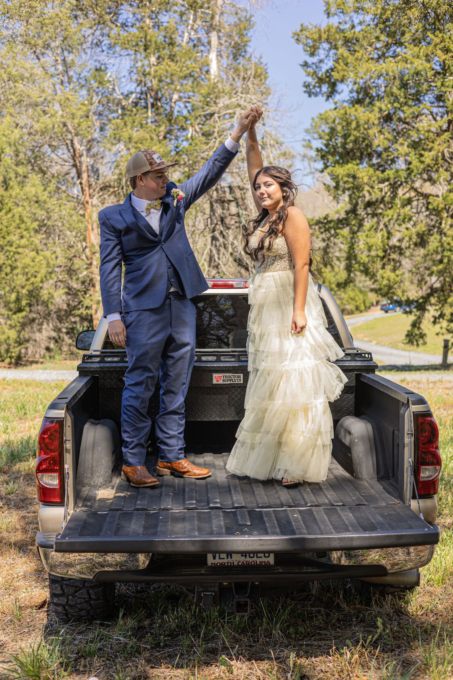 A bride and groom dance on a pickup truck bed outdoors, wearing wedding attire with cowboy boots and hat.