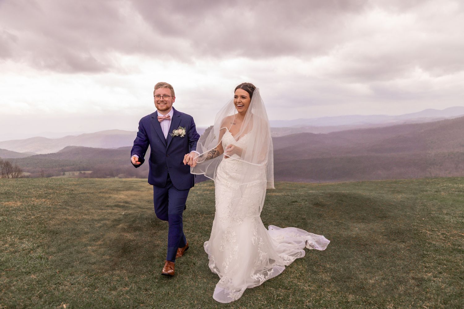 Newlyweds walking together on a grassy hillside with mountains in the background.
