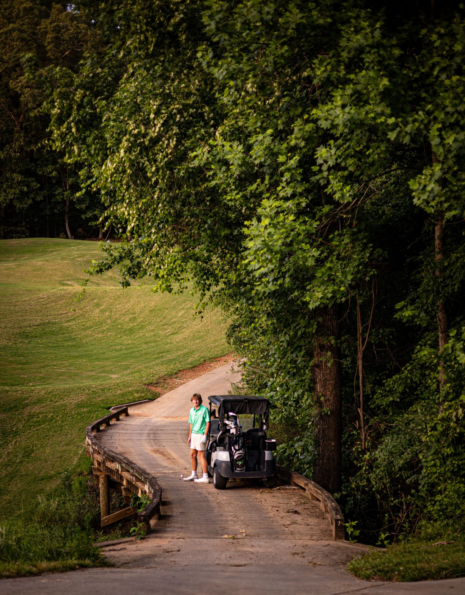 Two golfers stand beside a golf cart on a winding path surrounded by lush green trees on a sunny golf course.