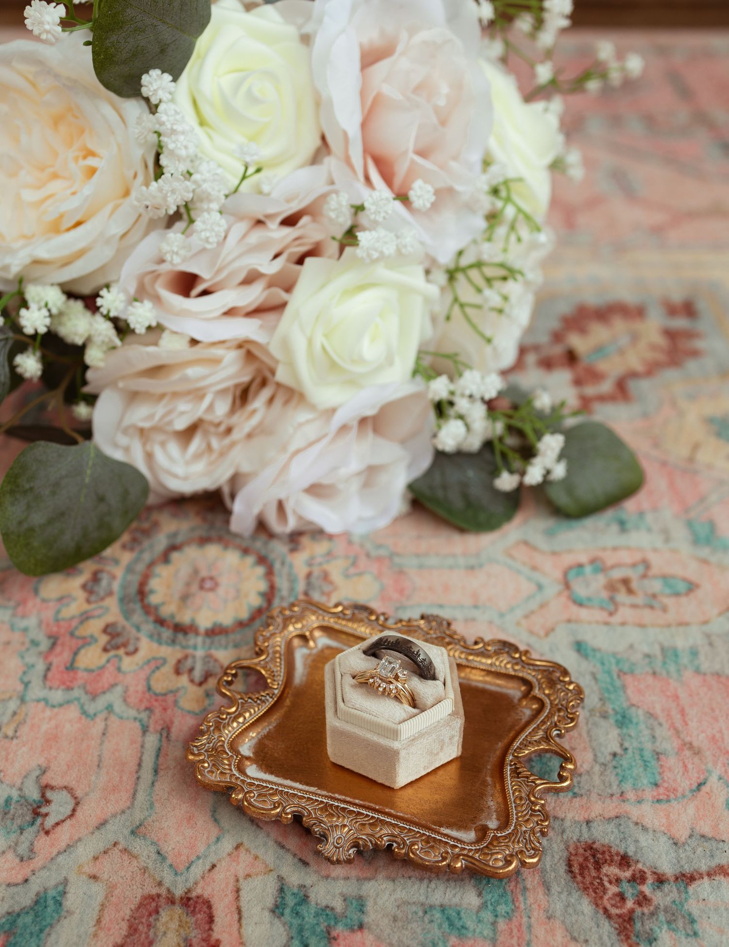 Elegant wedding flat lay with blush roses bouquet and gold ring box on ornate tray atop a floral rug.