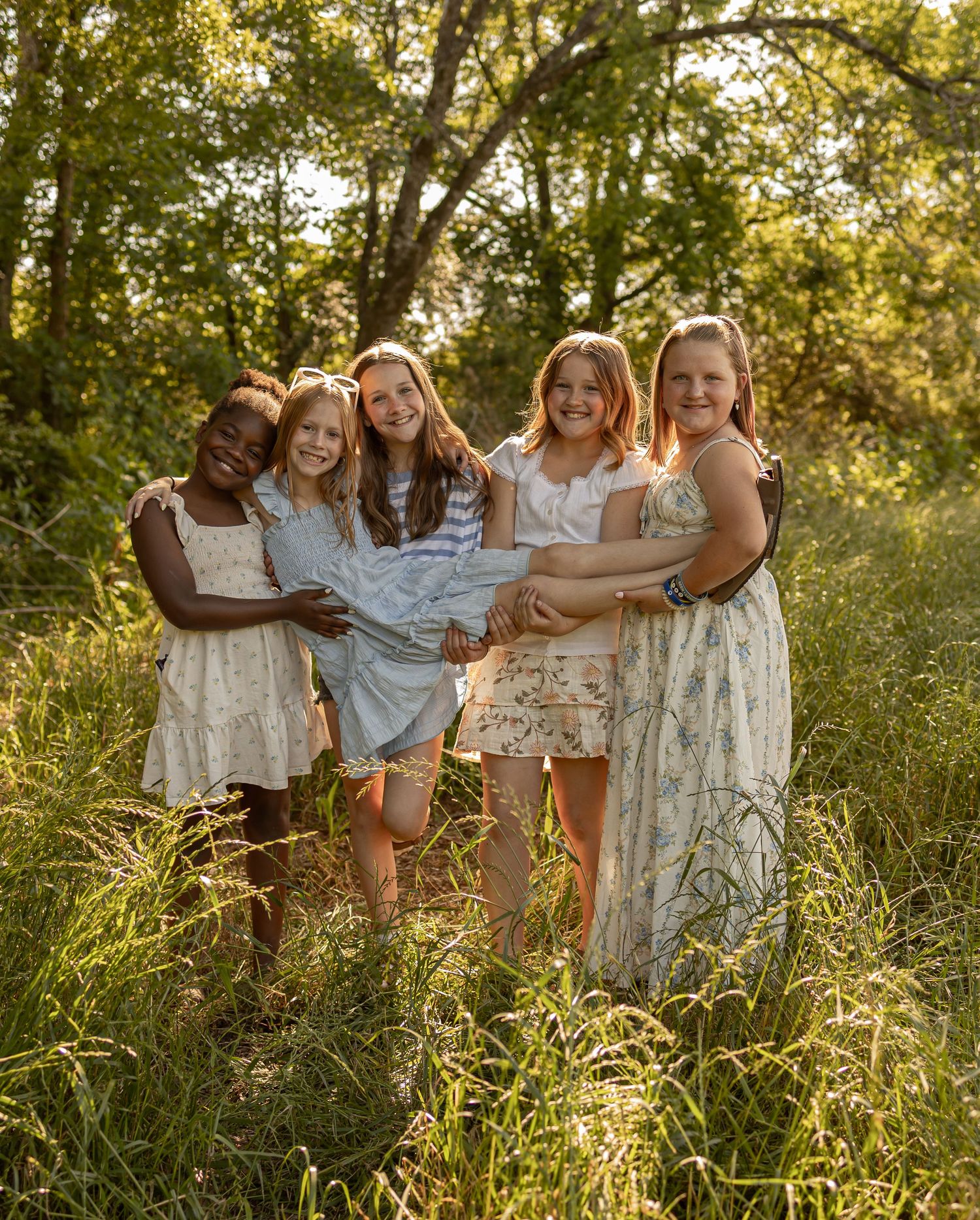 Five girls in floral dresses laughing together in a sunlit meadow surrounded by lush green trees.