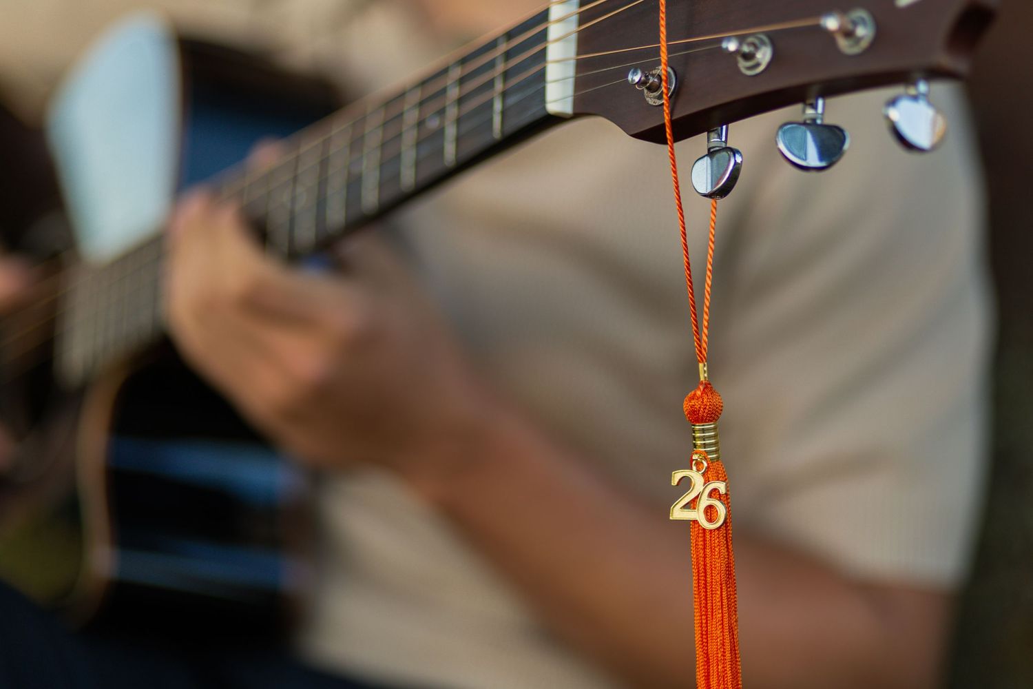 Close-up of guitar neck with orange tassel charm showing number 26, musician strumming in soft background.