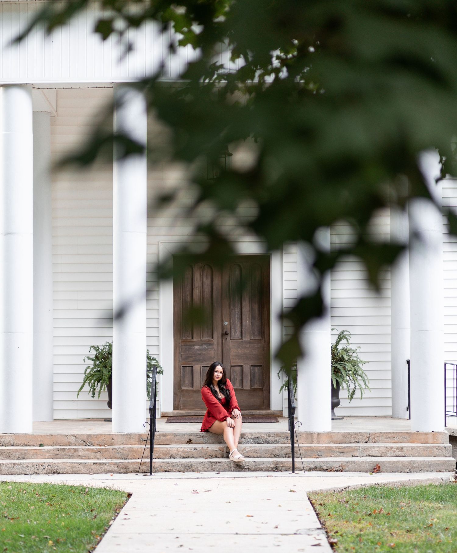 A person in a red shirt walks along a concrete path leading to a white building with columns and wooden doors.