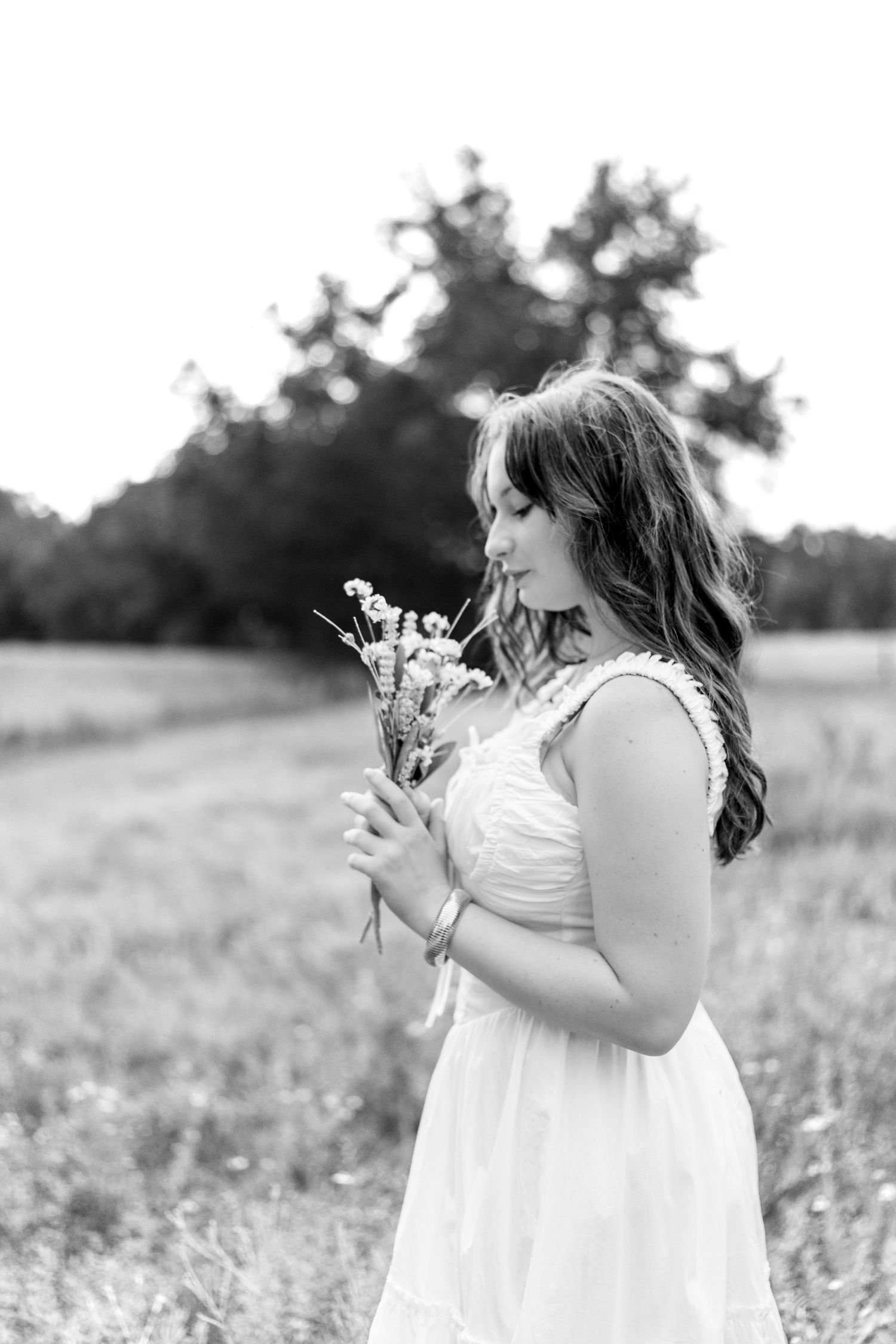 A woman in a white dress stands in a meadow holding wildflowers while looking down in black and white photography.