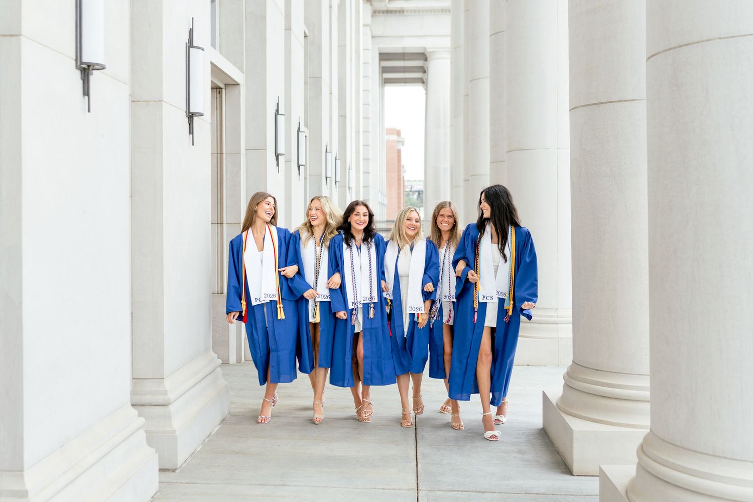Six women in blue graduation gowns walk together between white columns, celebrating their academic achievement.