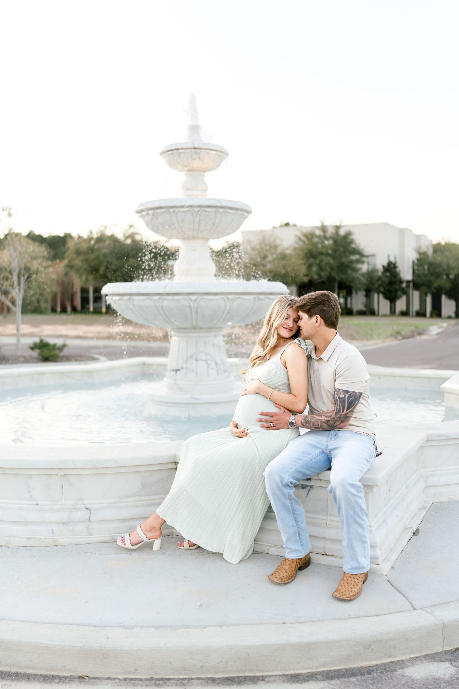 Couple sitting on a white tiered fountain, man kissing pregnant woman's cheek while holding her baby bump.