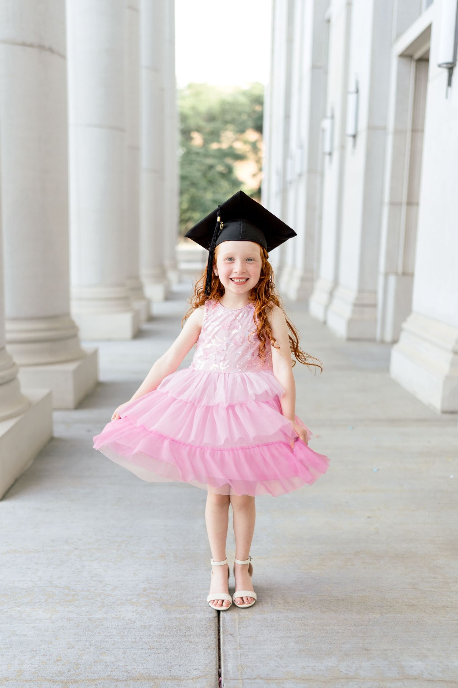 Young girl in pink tulle dress and graduation cap smiling outdoors near white columns.