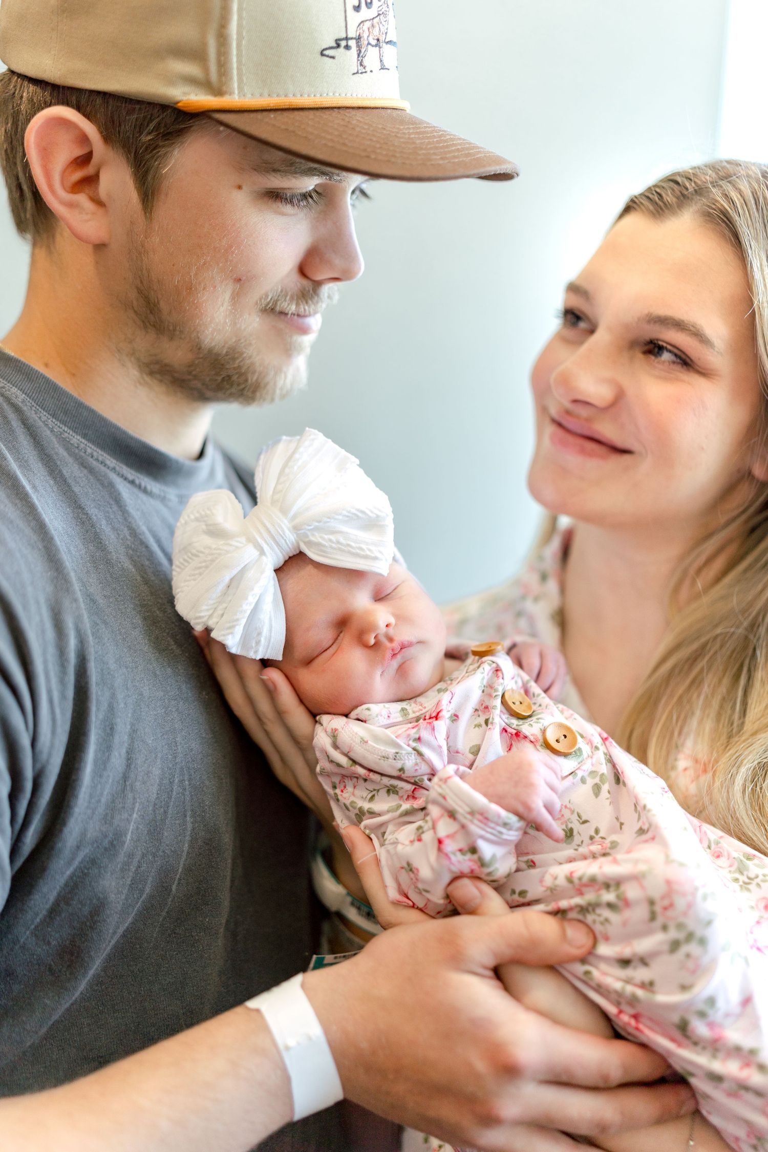Happy young couple smiling at their sleeping newborn baby girl wrapped in a floral swaddle with a bow headband.