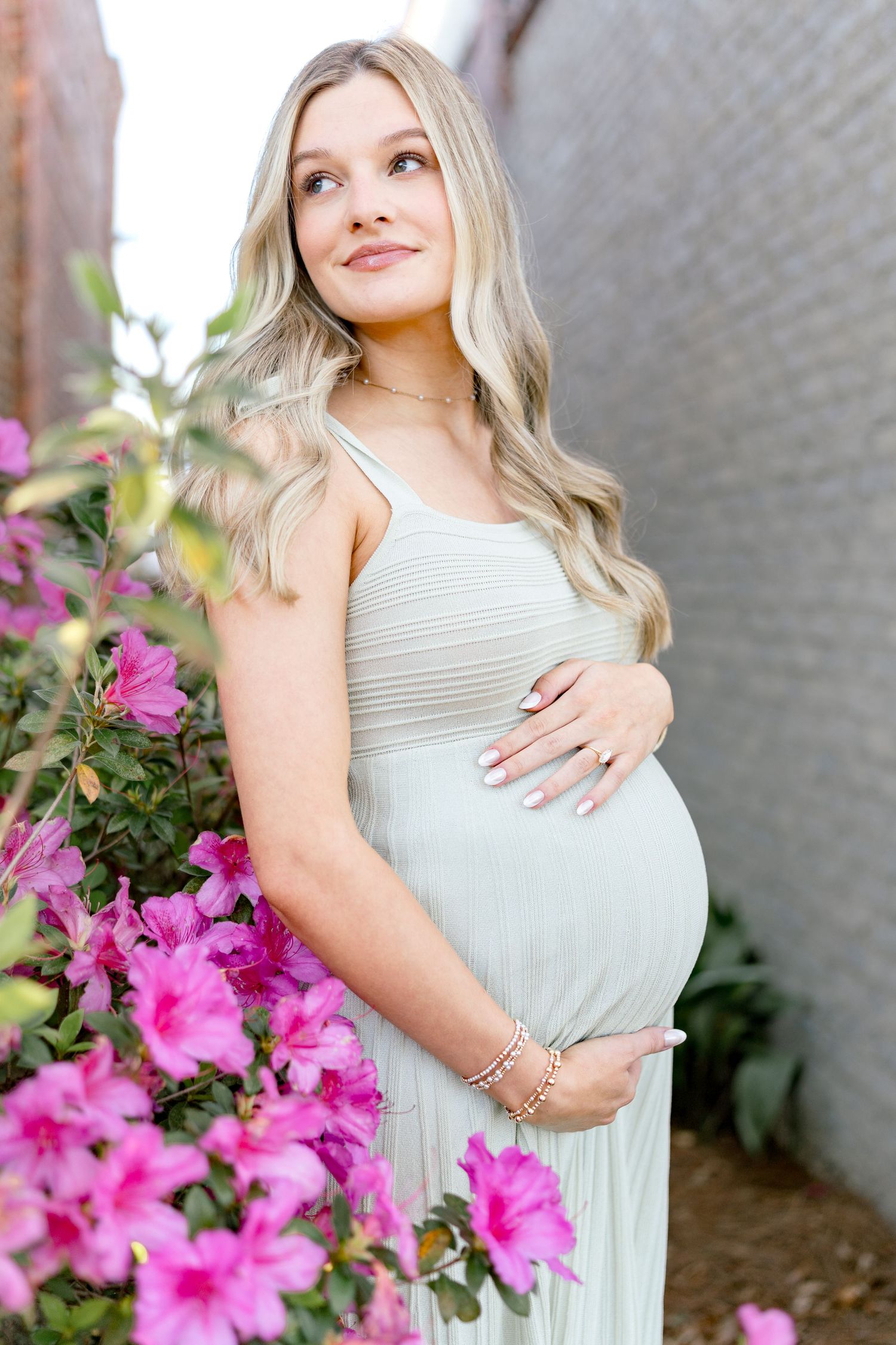 Pregnant woman in sage green dress cradling baby bump beside pink azalea blooms in outdoor maternity photo.