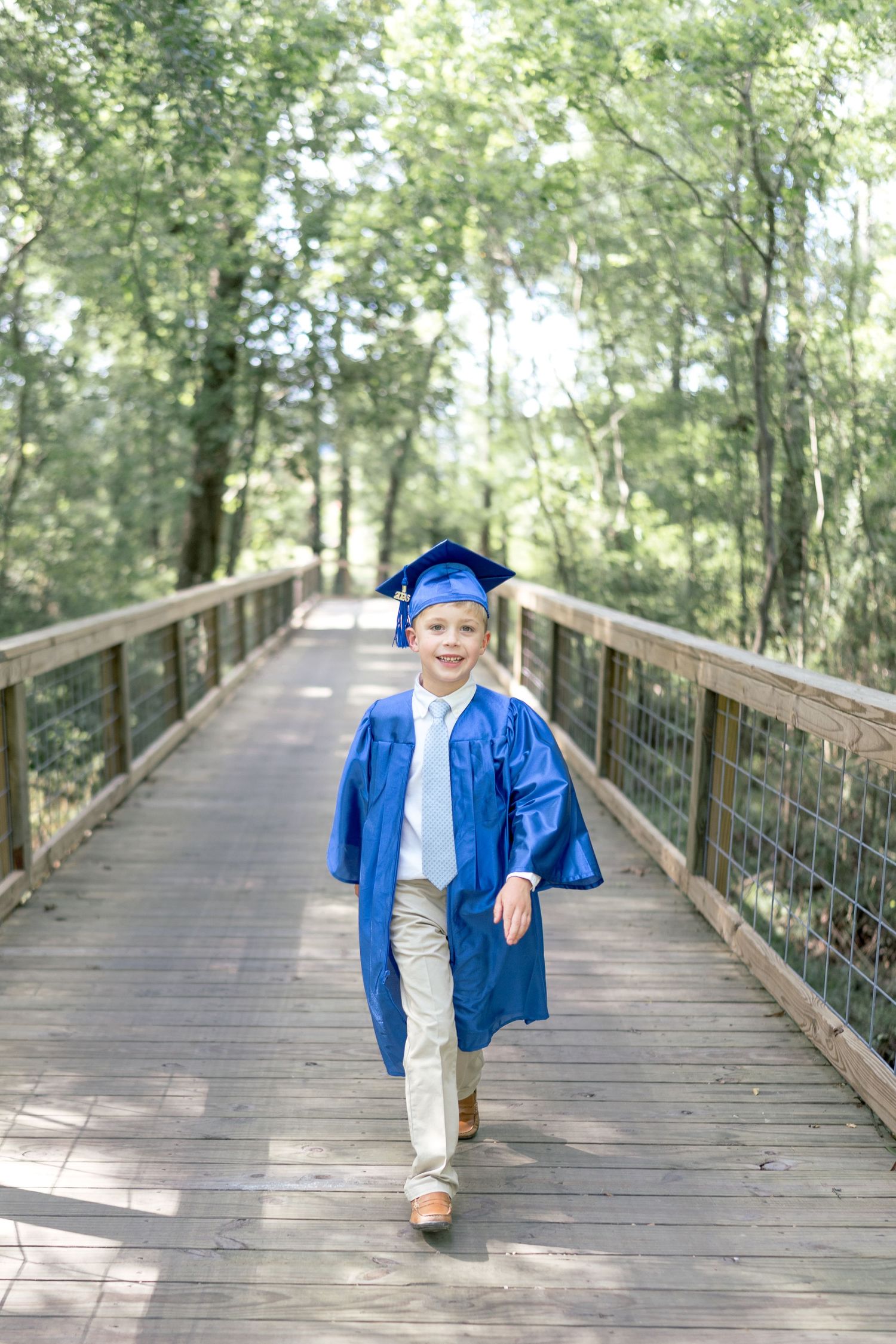 Young child in blue graduation cap and gown walking on wooden bridge surrounded by green trees.