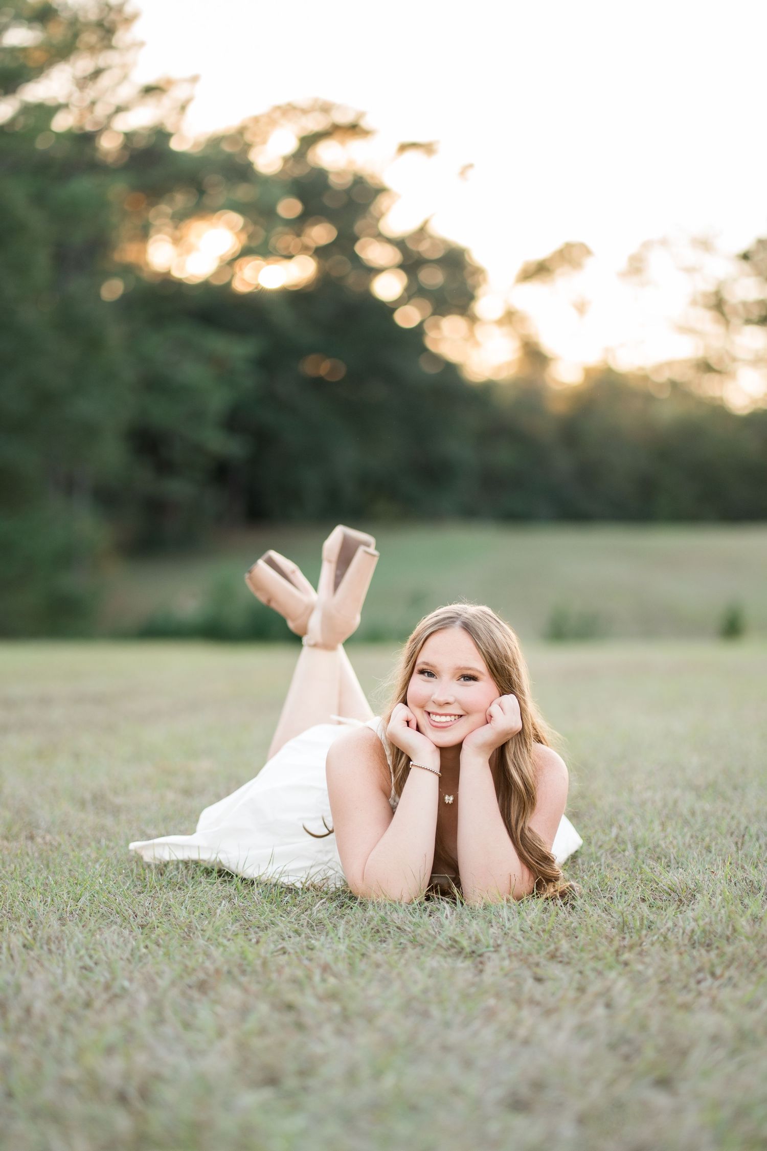 Smiling young woman in a white dress lying on her stomach in a grassy field, chin resting on her hands, with golden sunset light behind.