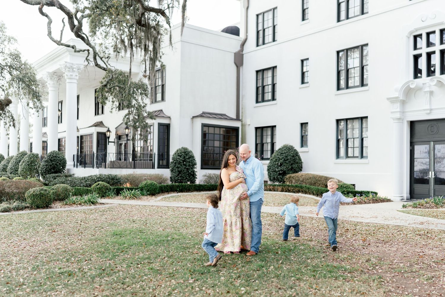 Family of five posing outdoors in front of a grand white building with lush greenery and Spanish moss-draped trees.