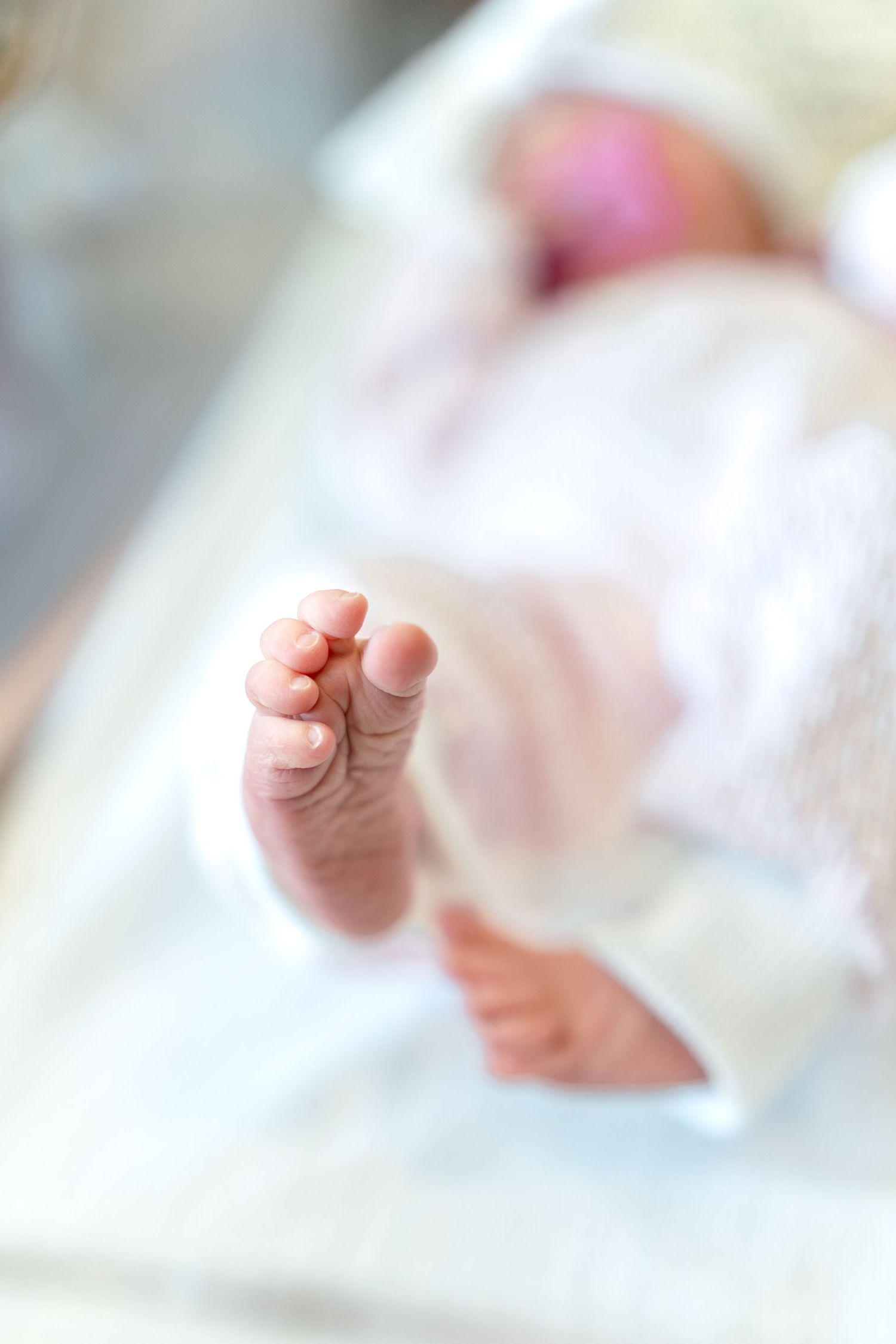 Close-up of tiny newborn baby feet in white clothing, with blurred infant lying in hospital bassinet background.