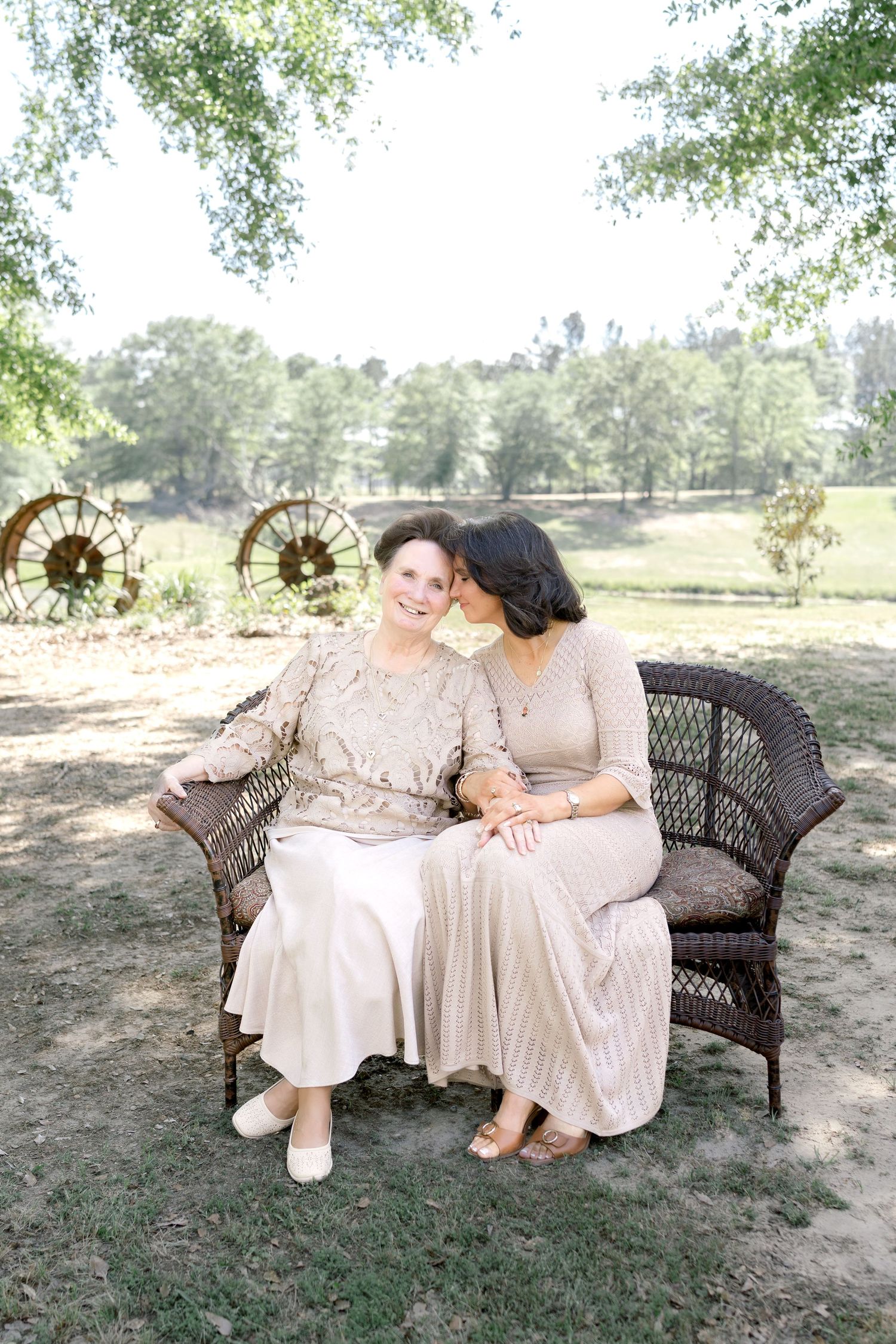 Two women in elegant neutral dresses sit closely on a wicker bench outdoors, sharing a tender moment in a lush garden setting.