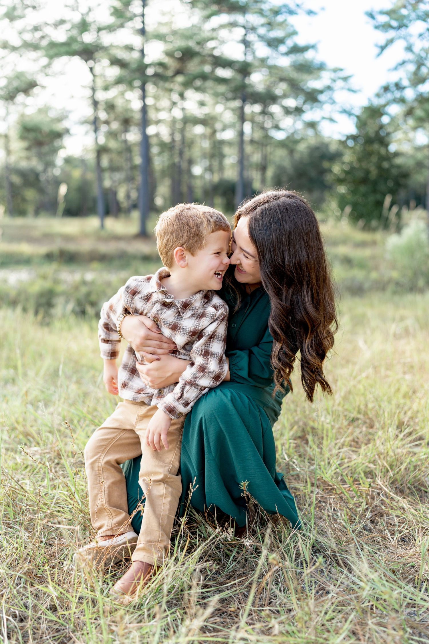 A tender moment shared outdoors in a grassy field with pine trees in the background on a sunny day.
