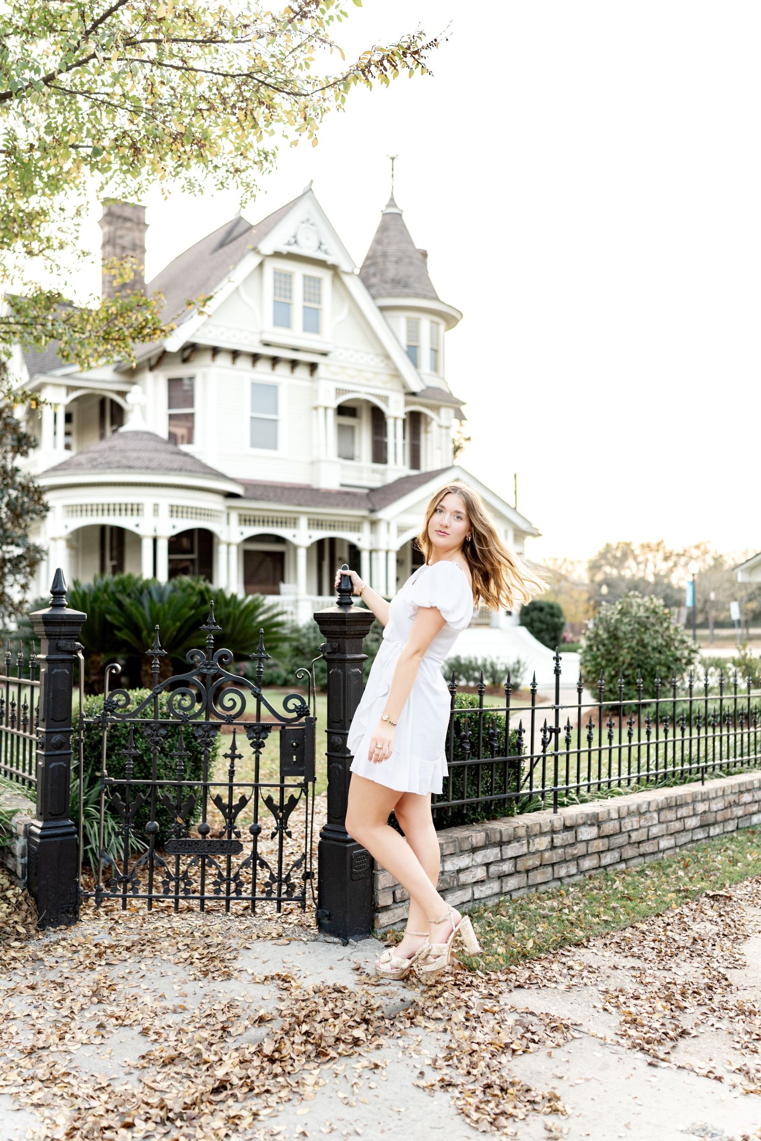 Young woman in white dress posing by ornate iron fence before a grand Victorian house with autumn leaves.