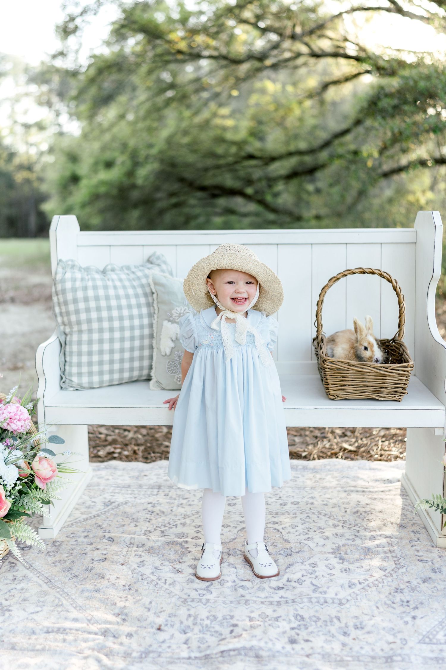 Toddler girl in light blue dress and straw hat smiling beside a wicker basket with a bunny on a white bench outdoors.