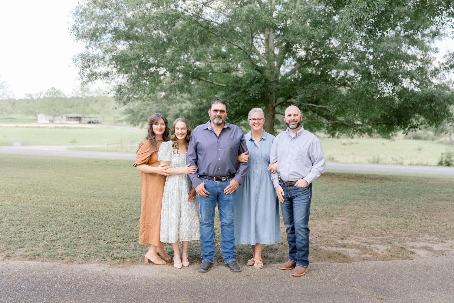 Family of five posing outdoors on a gravel path surrounded by lush green trees during a portrait session.