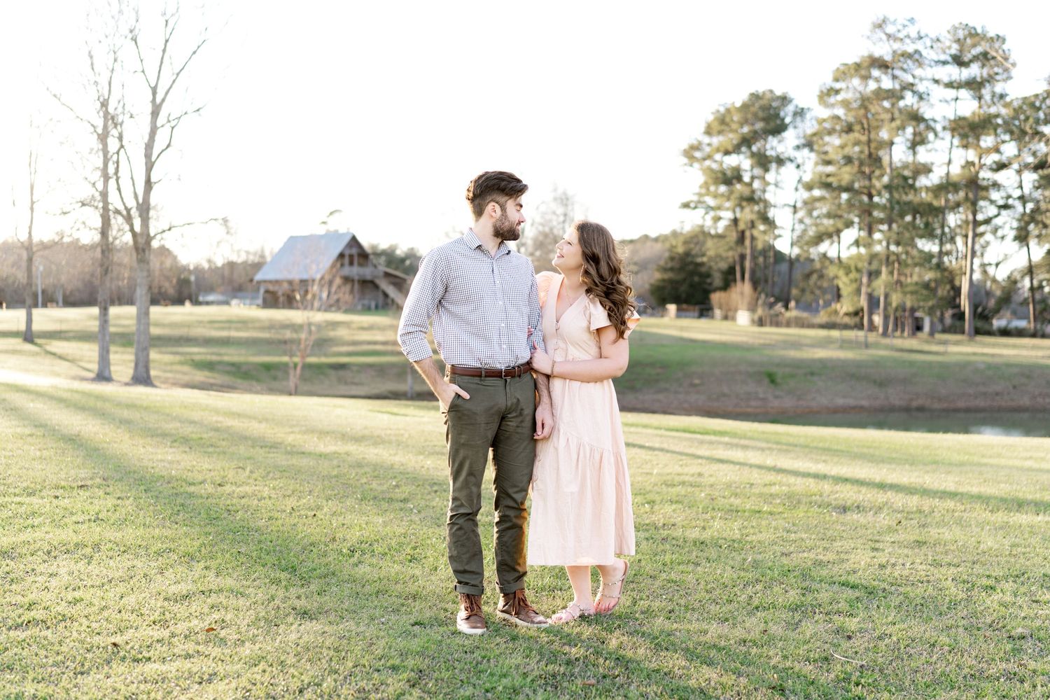 Couple standing together on a green lawn at golden hour with rustic barn and trees in background.