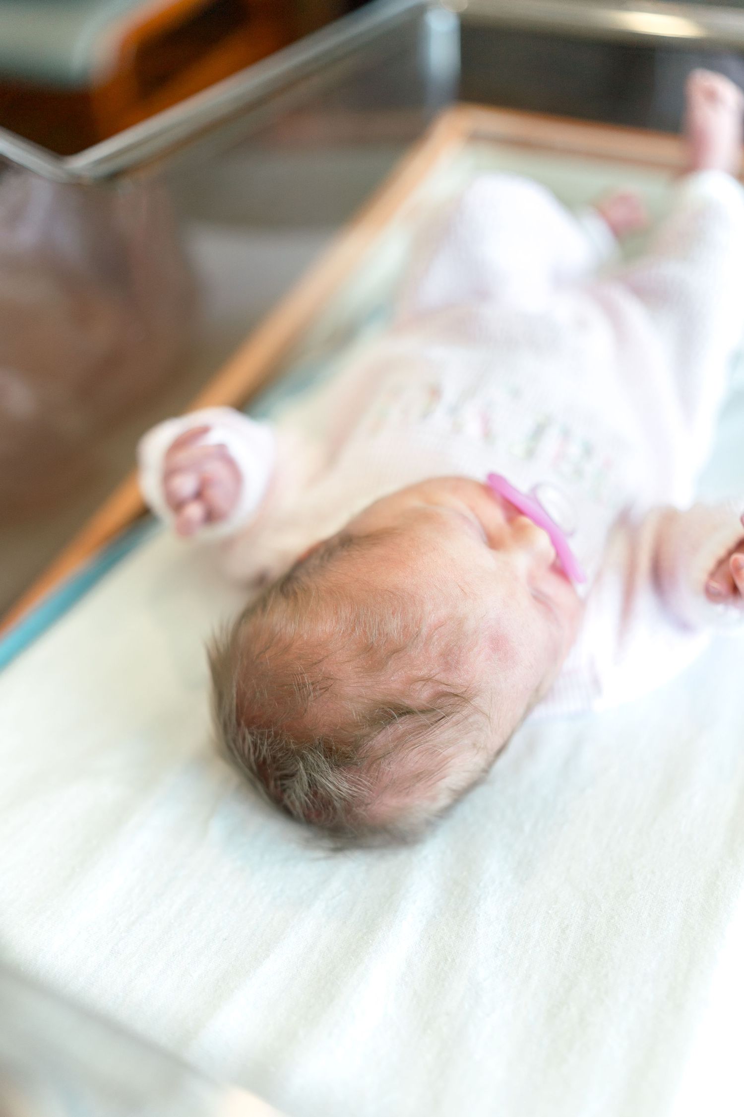 Newborn baby girl in hospital bassinet wearing pink headband, lying on white blanket in clear plastic crib.