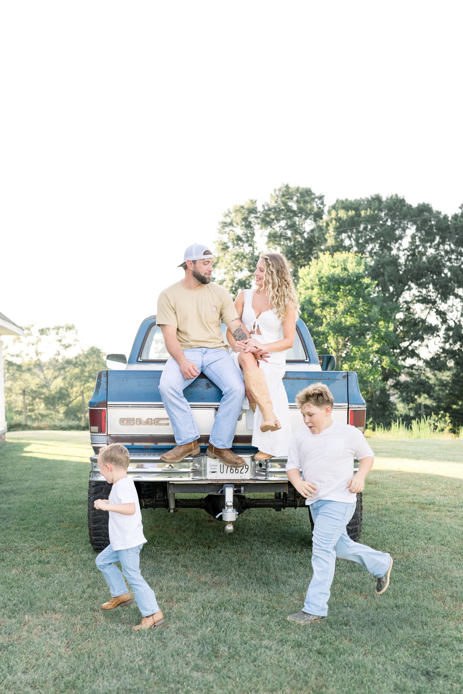 Family of four enjoying outdoors by a GMC truck, parents sitting on tailgate while two boys play in the grass.