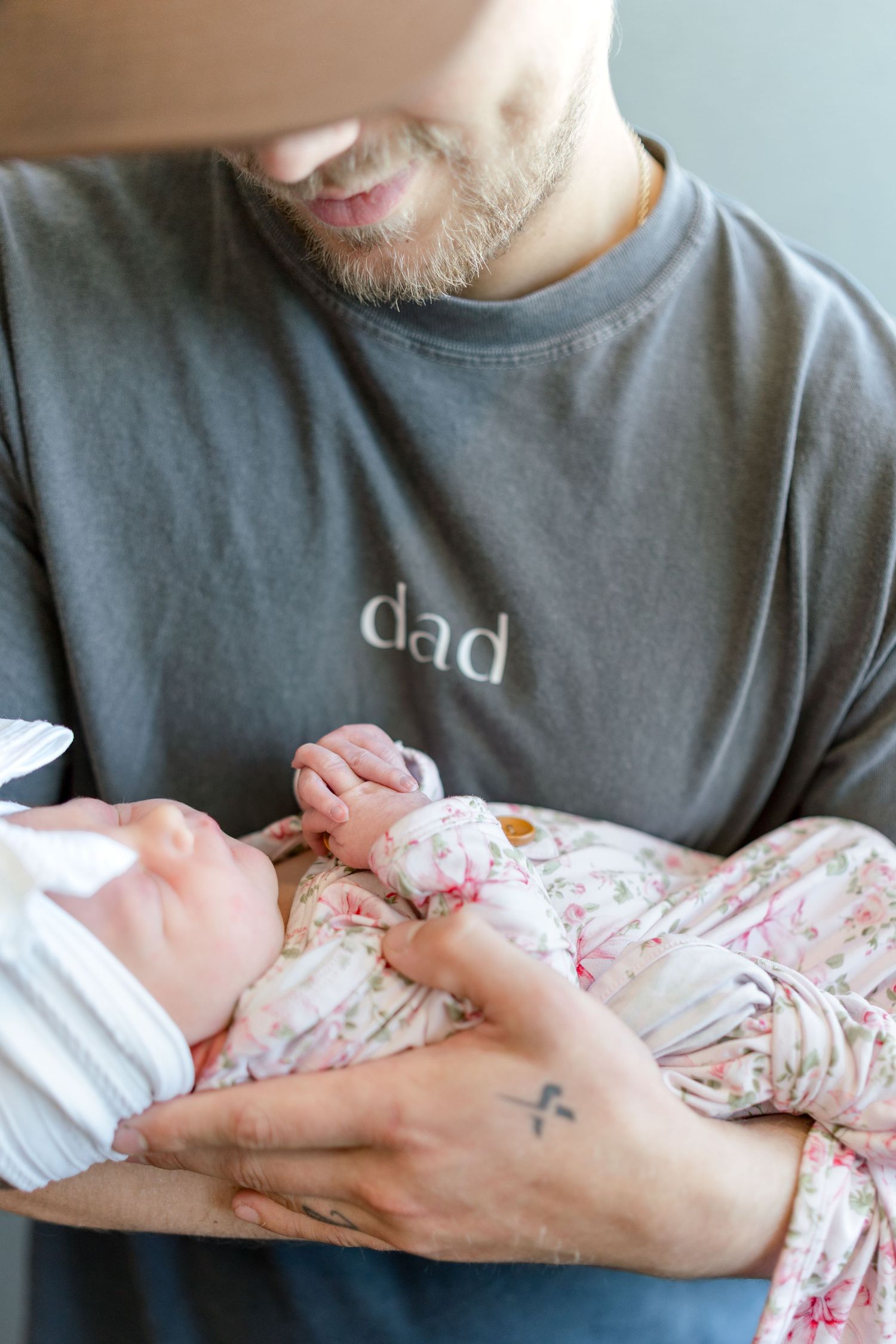 A bearded dad in a gray 'dad' shirt tenderly holds his newborn baby dressed in a floral outfit.