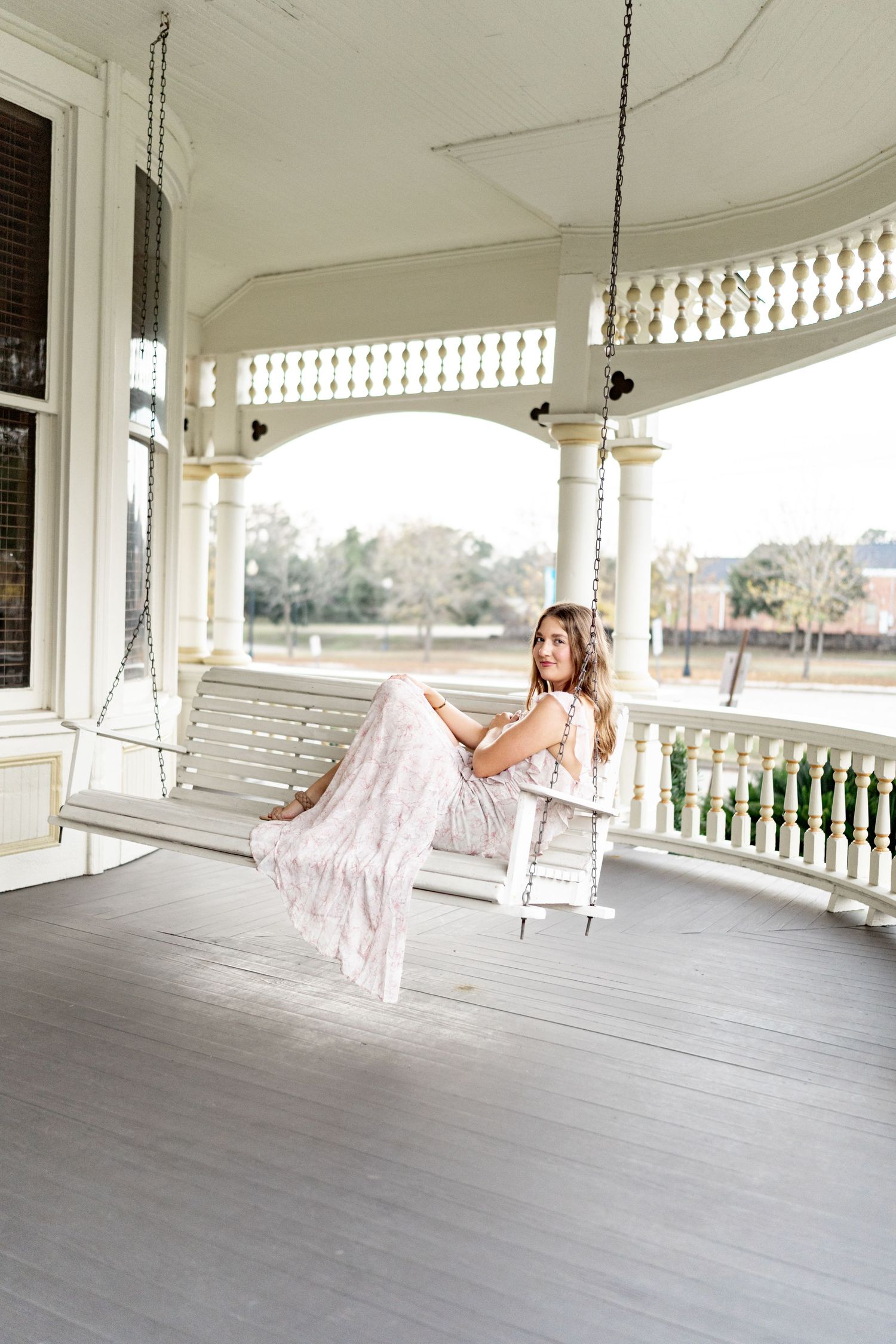 A person in a flowing pink dress relaxes on a white porch swing at an elegant Southern-style veranda.