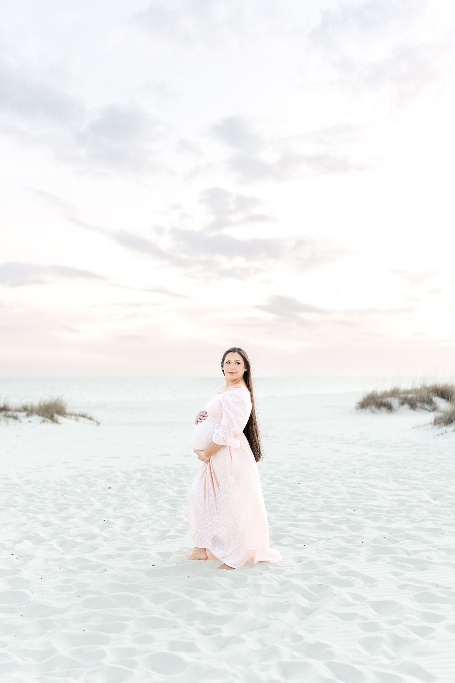 Pregnant woman in pink gown on white sand beach at sunset, cradling her baby bump.