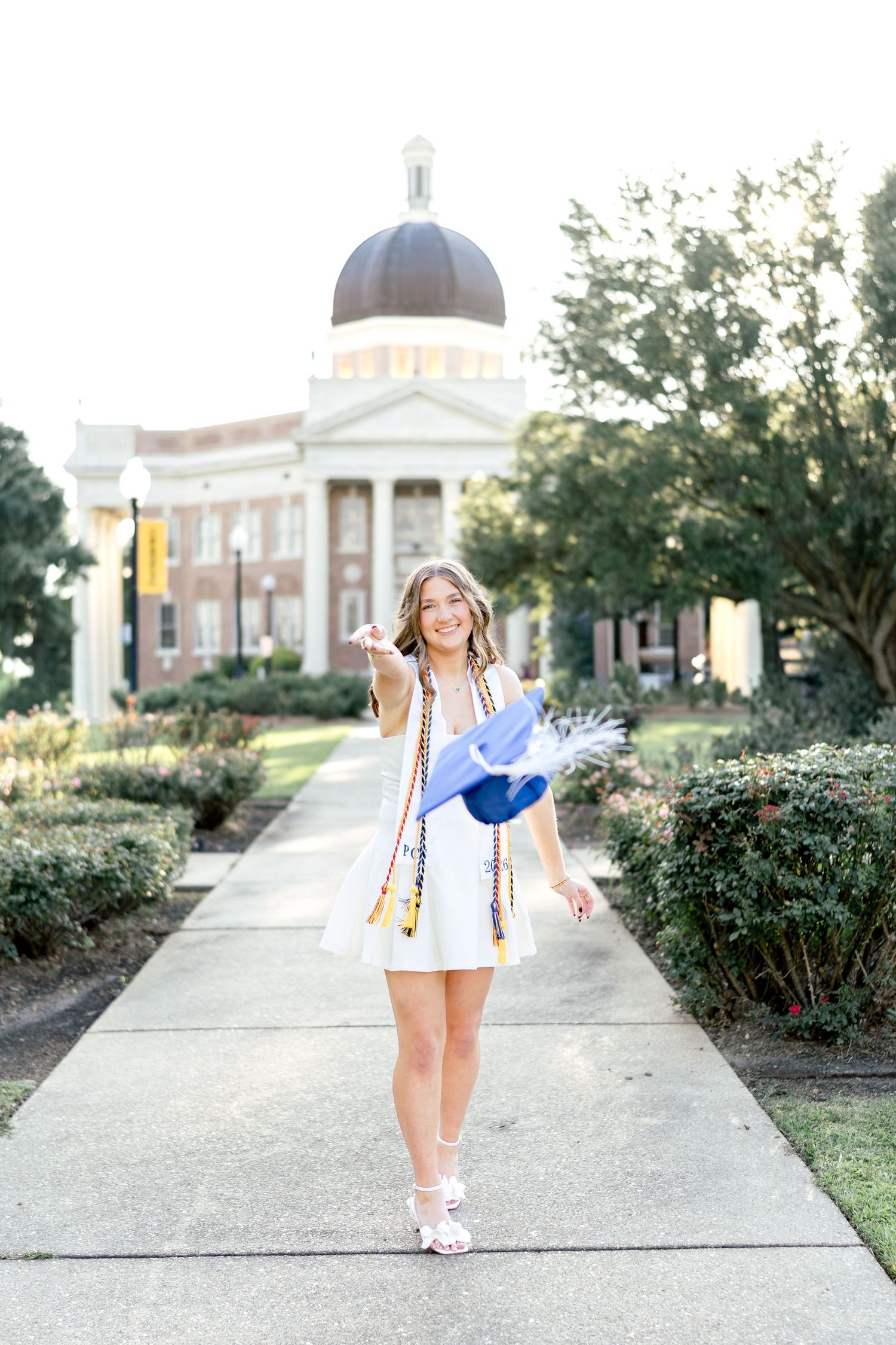 Smiling graduate in white dress holds blue cap on campus walkway with domed building backdrop.