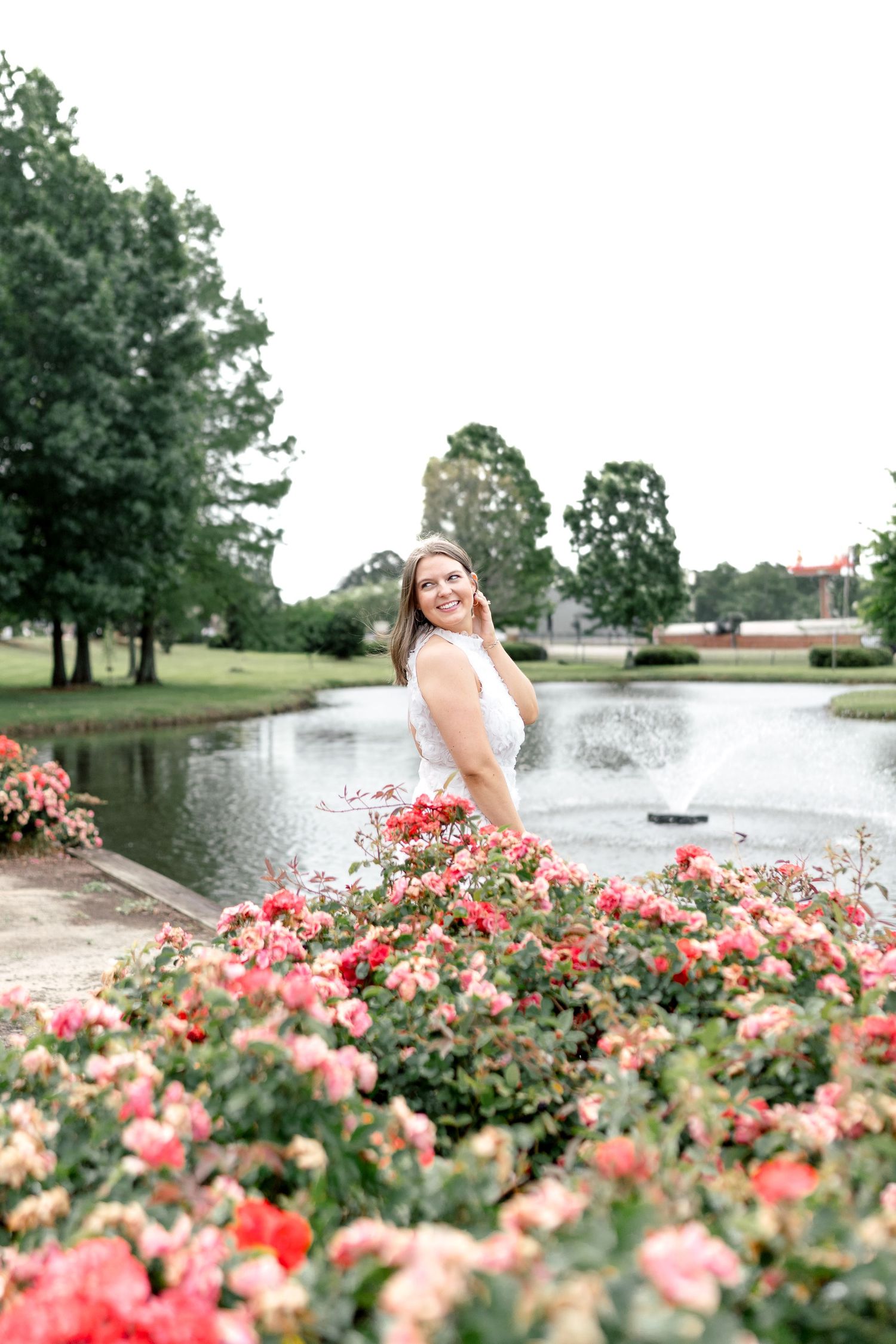 A smiling woman in a white dress poses among vibrant pink roses near a serene pond in a lush green park.